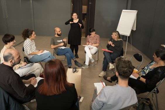 Περίπου 10 άτομα σε ανοιξιάτικα ρούχα κάθονται σε κύκλο σε ένα δωμάτιο με γκρίζους τοίχους. Ακούν μια ξανθιά γυναίκα σε γκρίζα ρούχα που μιλάει. Κοντά, μια γυναίκα σε μαύρα ρούχα διερμηνεύει στη νοηματική γλώσσα.