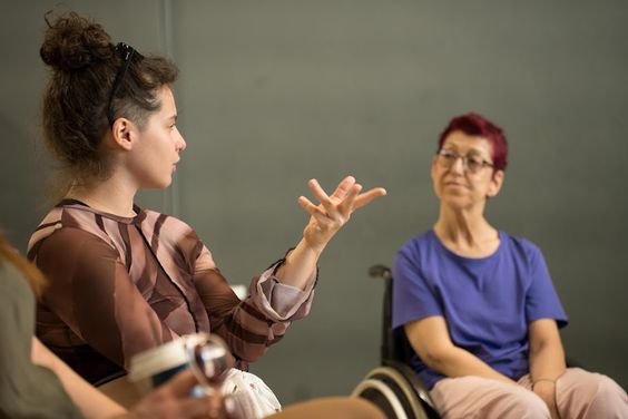 In a room with grey walls, a young woman with curly hair tied up in a bun and wearing a brown top appears to be explaining something, gesturing with her hands to people outside the frame. Next to her, a red-haired woman in a wheelchair, wearing glasses, watches her attentively.